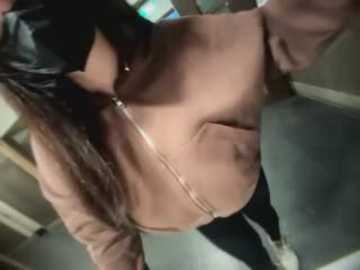 Media: A video of a woman with light skin and long black hair, wearing a pink hoodie with a zipper, black choker, and black pants. She is standing in a dimly lit room with a wooden door and a blurred background.
