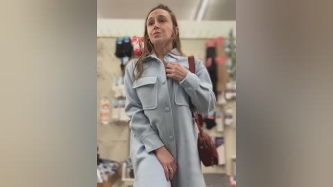 Video of a blonde woman in a light blue coat and maroon handbag, taking a selfie in a brightly lit store.