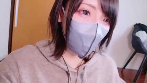 Video of a young East Asian woman with short, straight black hair, wearing a light grey face mask and beige hoodie, indoors against a wooden door and chair.