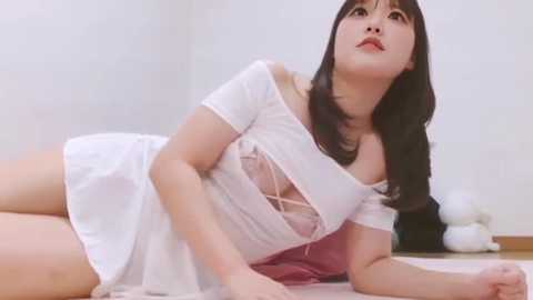 Video of a young Asian woman with long black hair and fair skin, lying on a bed in white lingerie with a revealing neckline, looking upwards with a serene expression.