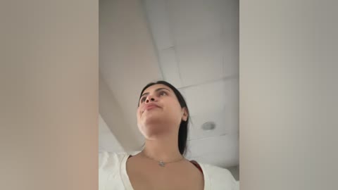 Video of a young woman with medium skin tone and dark hair, wearing a white top, looking upwards with a thoughtful expression in a modern, minimalist room.