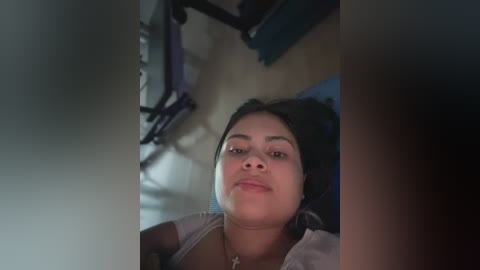 Video of a smiling, light-skinned woman with medium-length black hair, wearing a black headband and a light-colored top, seated on a wooden floor. A metal folding chair and a blue object are visible in the background.