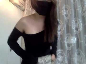 Video of a light-skinned woman with long brown hair, wearing a black off-the-shoulder sweater and a face mask, standing in a bathroom with a white shower curtain featuring floral patterns.