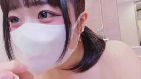 Video of an Asian woman with fair skin, wearing a white face mask, black hair, and a strapless top. She's indoors, with a blurred background showing white walls.