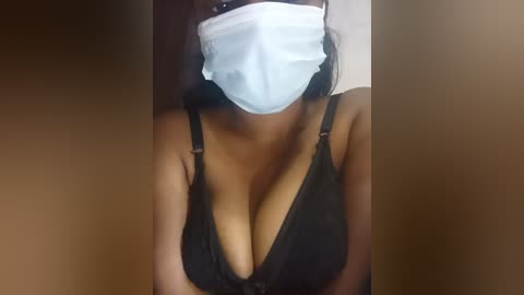 Video of a woman with a medium skin tone, wearing a black lace bra that accentuates her large breasts, a white surgical mask covering her mouth and nose, and long dark hair.