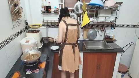 Video of a woman in a beige apron cooking at a kitchen counter with white cabinets and a black countertop. The background includes a sink, utensils, and dishes.