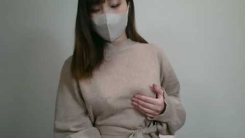 Video of a fair-skinned Asian woman with straight, shoulder-length brown hair, wearing a beige turtleneck sweater, gray face mask, and a light gray tie, standing against a plain, light gray background.