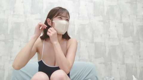 Video of a young Asian woman with shoulder-length brown hair, wearing a white face mask, adjusting her black camisole strap, seated on a blue couch against a textured, light-colored wall.