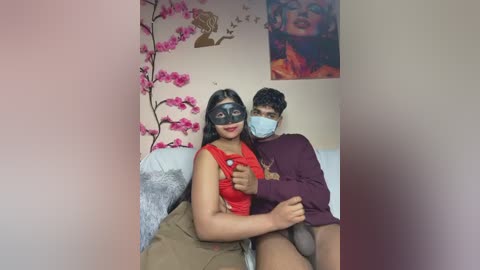Video of a South Asian couple in a cozy bedroom, wearing masks and red clothes, with floral decor and a Buddha painting on the wall.