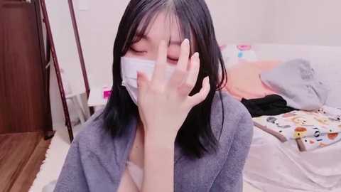 Video of an Asian woman with straight black hair, wearing a gray robe, covering her face with a white mask, in a messy bedroom with a bed and a wooden floor.