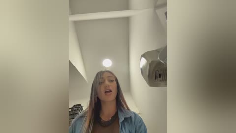 A video of a young woman with long, straight brown hair, wearing a blue denim jacket and a black top, standing in a modern, well-lit room with white walls and a metallic ceiling fixture.