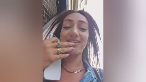 A candid video of a young woman with light skin and shoulder-length auburn hair, wearing a denim jacket and a silver necklace, smiling while touching her mouth with a ring-adorned hand.
