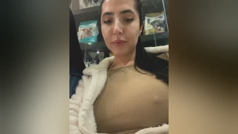 Video of a young woman with medium skin tone, dark hair, and brown eyes, wearing a beige ribbed top and a white fluffy jacket, sitting in a pet store, surrounded by pet food displays.