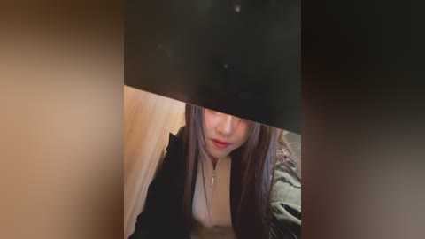 Video of an Asian woman with long brown hair, wearing a black jacket over a white top, partially obscured by a black object. She appears to be in a dimly lit room with wooden and fabric textures visible.