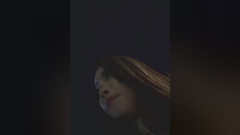 A dimly lit video of a young woman with long, straight brown hair, light skin, and dark eyes, gazing upward, set against a dark, shadowy background.