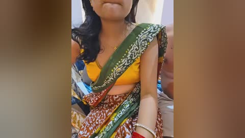 Video of a South Asian woman in a vibrant yellow blouse and multi-colored sari, with a gold necklace, sitting indoors, smiling.