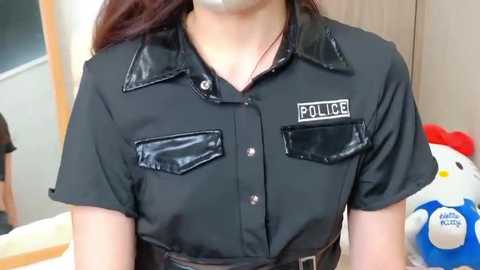 Video of a woman wearing a black, shiny police-themed short-sleeved shirt with a silver badge on the left chest pocket. Background features a wooden door and a white Hello Kitty plush toy.