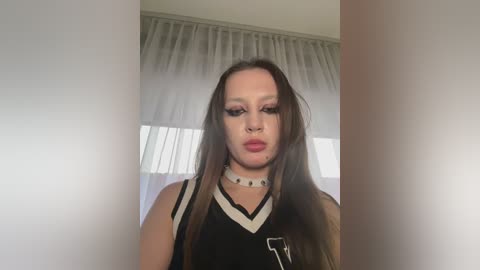 A video of a young woman with long brown hair, fair skin, wearing a black sports jersey with a white V-neck and a white choker. She has heavy eyeliner and red lipstick. Background shows semi-transparent white curtains.