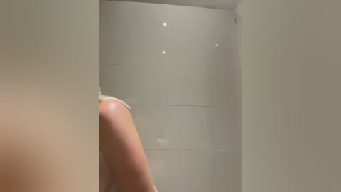 Video of a close-up view of a nude, light-skinned person's back against a plain, white-walled bathroom with a small, circular mirror reflecting light.