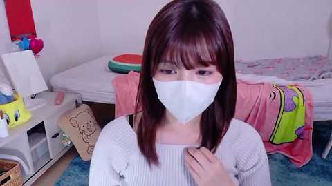 Video of a young woman with straight brown hair, wearing a white face mask, light grey sweater, and blue jeans, sitting on a bed with a pink blanket and cartoon character print. Background includes a nightstand with various items, a red wall, and a wicker basket.
