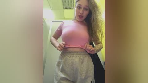 A video of a young, light-skinned woman with long, blonde hair, wearing a tight pink crop top and loose, grey sweatpants. She's pulling up her top, revealing a tattooed arm, in a dimly lit, narrow hallway.