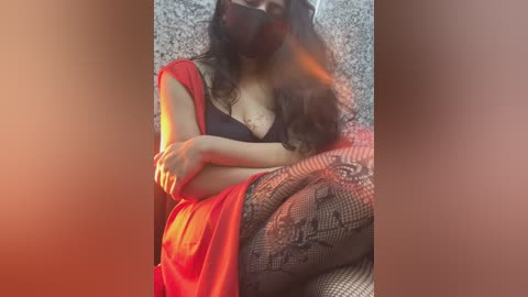 A video of a woman with medium skin tone and long dark hair, wearing a black bra, red sari, and black lace stockings, sitting on a textured surface with a warm, reddish-orange glow.