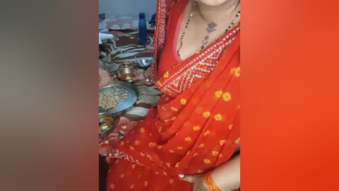 Video of a woman in a red sari with yellow floral embroidery, wearing a gold necklace, standing beside a cluttered kitchen counter with utensils and spices.
