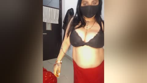 Video of a pregnant woman with medium skin tone, wearing a black bra and red skirt, standing indoors with a mask covering her mouth.