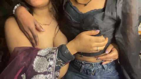 Video of two women, one with medium skin and long dark hair, wearing a sheer top and denim shorts, embracing a woman with a lighter skin tone and long, dark hair, wearing a lace bralette.