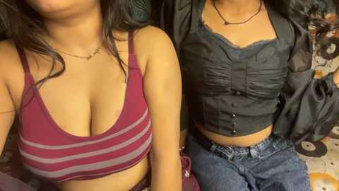 Video of two young women with medium skin tones, one wearing a maroon tank top with horizontal stripes, the other a black ruffled top, both seated on a patterned couch.