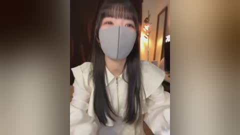 Video of an Asian woman with long black hair and a gray mask, wearing a white, frilly blouse, in a warmly-lit, indoor setting with blurred background.