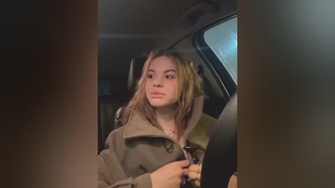 Video of a young woman with long, light brown hair, wearing a beige coat, driving in a dimly lit car at night.