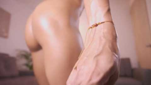 Video of a nude woman with light skin, her backside and foot in focus, wearing a gold anklet, blurred background of a beige room with a couch.