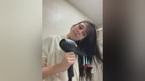Video of a young woman with long, wet black hair, wearing a white towel, using a black hairdryer in a tiled bathroom.