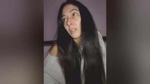 A video of a young Asian woman with long, straight black hair, wearing a grey off-shoulder sweater, and a neutral expression. Background is blurred, featuring a light purple wall.