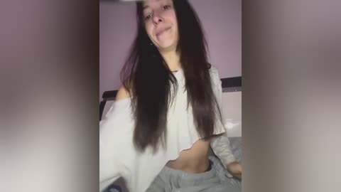 A young woman with long, straight dark hair, light skin, and a slim build, wearing a white off-shoulder crop top and gray sweatpants, sits on a bed in a dimly lit room.