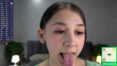 A close-up video of an Asian woman with dark hair, wearing a nose ring, and sticking her tongue out, in a dimly lit bedroom with a digital clock and plants in the background.