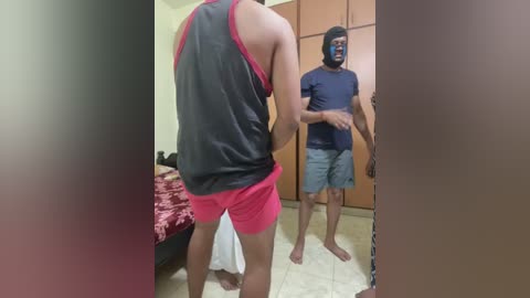 Video of a man in a black tank top and pink shorts standing near a man in a blue shirt and shorts, both facing a closed wooden wardrobe in a beige-tiled room.