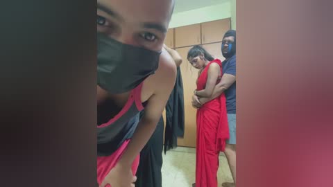 Video: Three people in a room, one with a mask, another in a red saree, and a man in blue shorts.