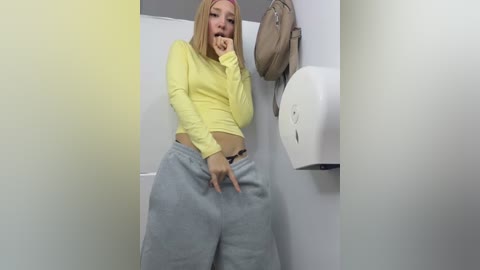 Video of a blonde, fair-skinned woman in a yellow crop top and loose grey sweatpants, standing in a public restroom. She has her hand on her chin and appears contemplative. A beige handbag hangs on the wall to her right.