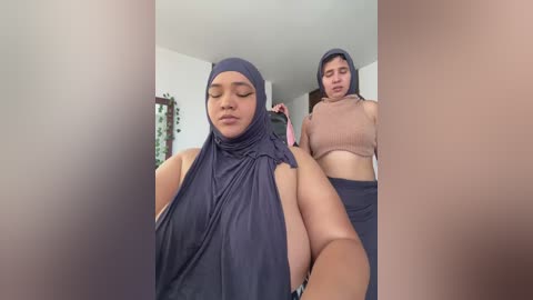 Video of a plus-sized woman with large breasts wearing a dark blue hijab and a beige crop top, standing next to a thin woman with medium breasts, both in a room with white walls.