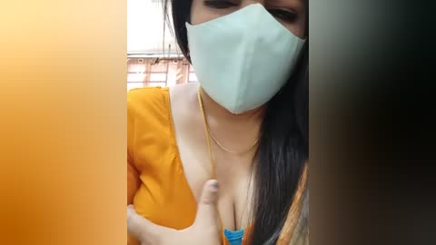 Video of a woman with long dark hair, wearing a yellow blouse, a blue bra, and a light blue mask, partially obscured by a black and orange gradient frame.