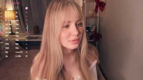 A video of a young Caucasian woman with long blonde hair, fair skin, and a soft smile, wearing a white top, indoors with a wooden floor and a red poinsettia plant in the background.