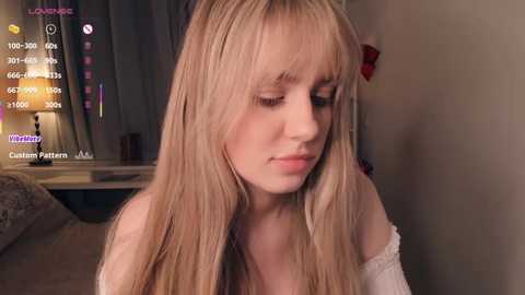 Video of a blonde woman with long hair, wearing a white off-shoulder top, sitting indoors with dim lighting, displaying a custom avatar in the background.