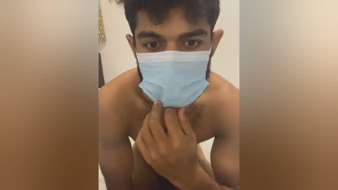 Video of a shirtless South Asian man with medium skin tone, dark hair, wearing a blue face mask. He holds the mask with his right hand, revealing a muscular chest. Background shows a beige wall and a partially visible door.