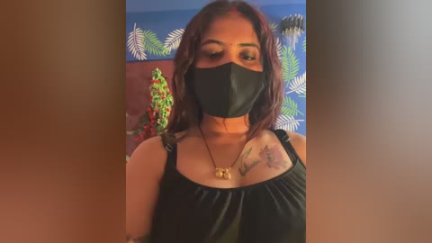 Video of a woman with medium skin tone, wearing a black mask, black top, and a necklace, standing against a blue and green floral wallpaper.