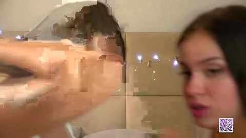 Video of a woman with long black hair and light skin, partially obscured by a transparent glass pane. She has a surprised expression, her eyes wide. Background shows a beige tiled wall.