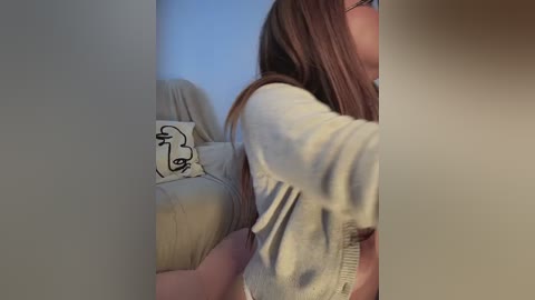 A video of a young woman with long brown hair, wearing a beige sweater, captured in a side profile. She's standing beside a beige couch with a white pillow featuring a minimalist face drawing. The background is a light-colored wall.
