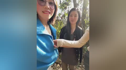 Video of two Asian women in a forest; one with glasses and a blue shirt, the other with a black jacket, holding hands.