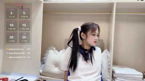 Video of an Asian girl with long black hair in a white school uniform, standing in a locker room with open wooden lockers and white towels.